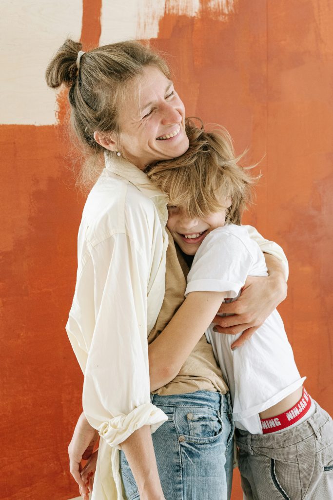 Happy mother embracing her son with a big smile, standing together in front of a painted orange wall.