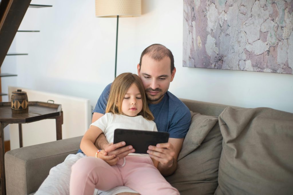 ather sitting on a sofa with his young daughter, both looking at a tablet together at home.
