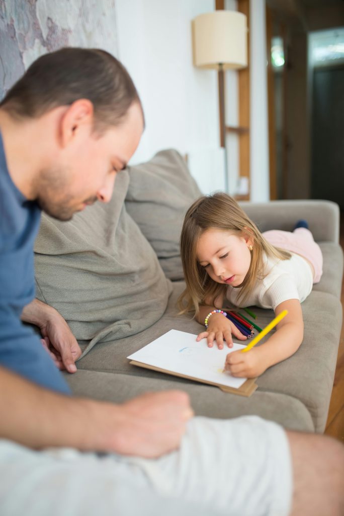 Young girl lying on a sofa drawing with coloured pencils while her father watches and supports her