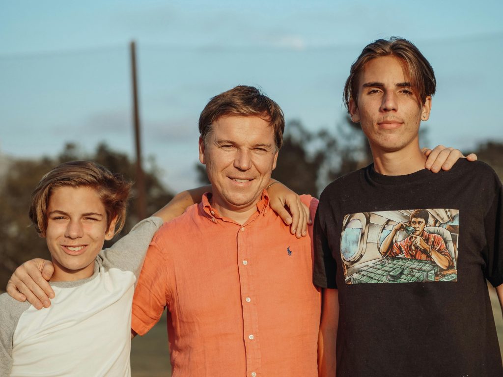 Father standing outdoors with his two teenage sons, smiling together with arms around each other.